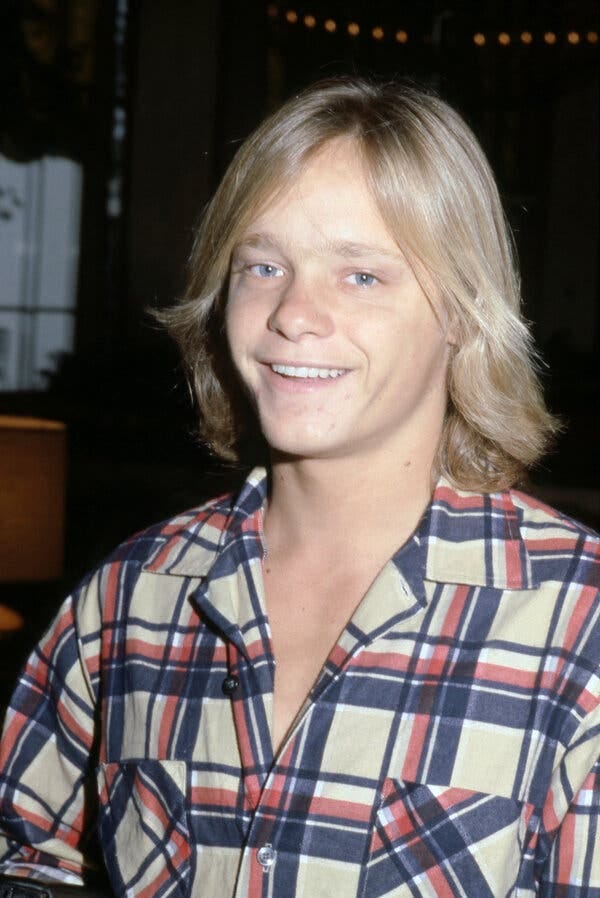 A smiling teenage boy with long blond hair in a plaid shirt.