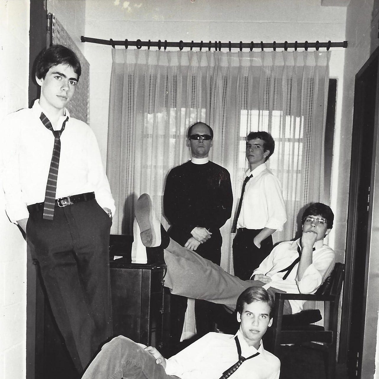 Four high school aged young men pose with a priest wearing sunglasses in a scene reminiscent of a first-wave band's album cover