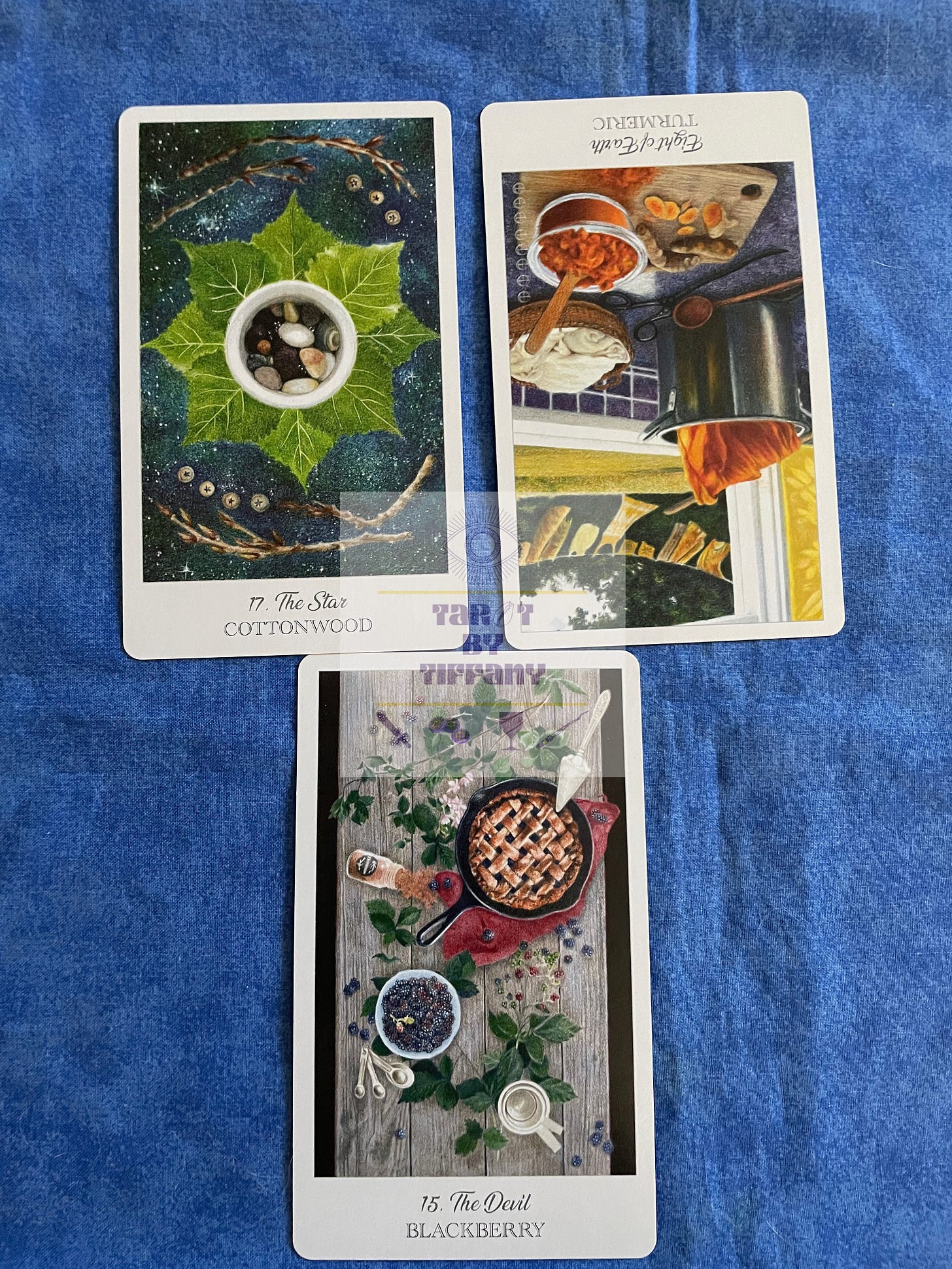 3-card reading with the Herbcrafter’s Tarot. The cards are laid on a cobalt blue cloth background. Top two cards (L-R): The Star (Cottonwood), Eight of Earth (Tumeric) reversed. Bottom card: The Devil (Blackberry).
