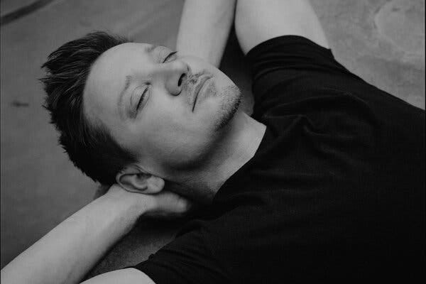 Fotografía en blanco y negro de Jeremy Renner tumbado con las manos detrás de la cabeza. Tiene los ojos cerrados y lleva una camiseta negra.