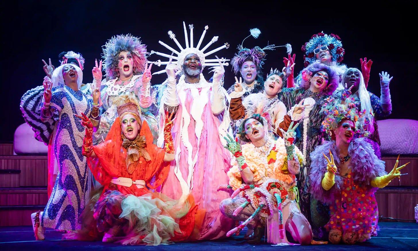 Le Gateau Chocolat, a black man wearing a spiked white headdress and a pink and white gown stands at the center of a colorful, multiracial cast of singers in extravagant costumes, including Taylor Mac. They are all holding up fingers in the air as they gesture and sing.