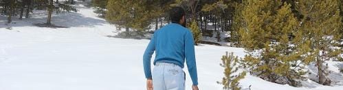 Jerome Ellis, wearing blue sweater and jeans, stands back to the viewer, in a clearing of snow with green trees in the background.