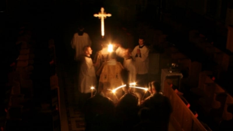 A photograph of an Easter Vigil, with candlelight illuminating the priest, acolytes, an cross.