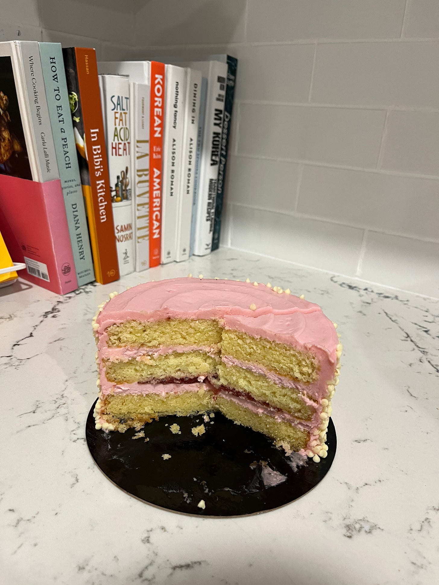 a circular pink frosted vanilla three-layer cake with half the cake already cut and eaten, sits atop a marble countertop with various cookbooks in the background.