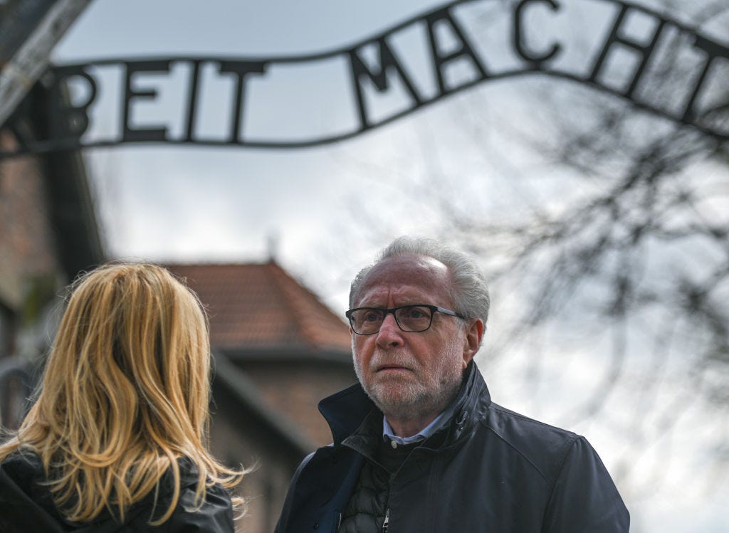 A man with white hair and close cropped beard in glasses and a dark coat stands under the sign to Auschwitz