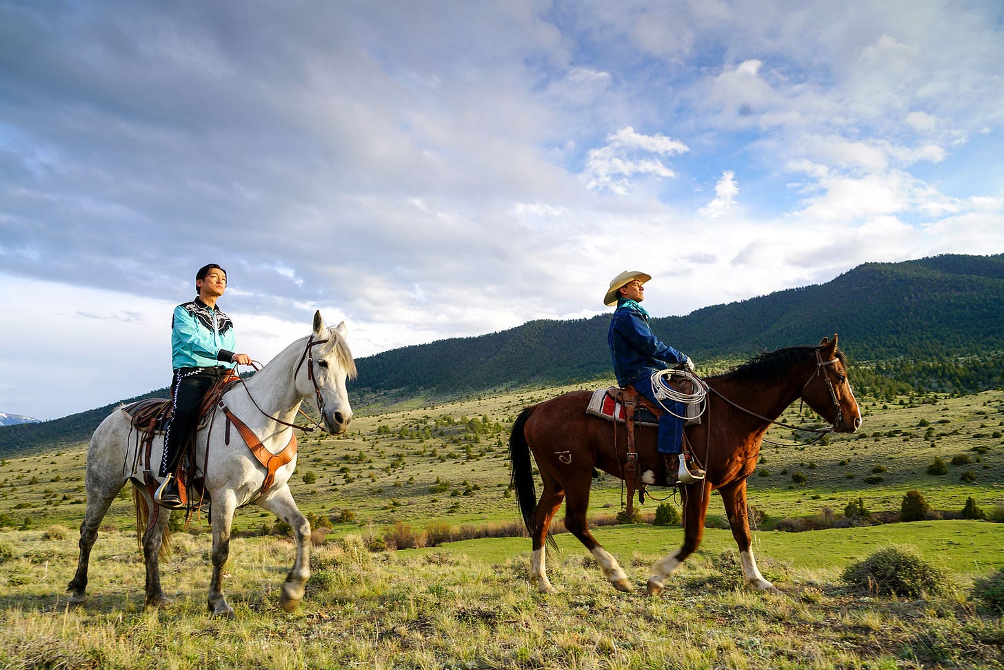 A Japanese man in colorful Western wear and a Hispanic man ride horses in a wide Montana landscape.