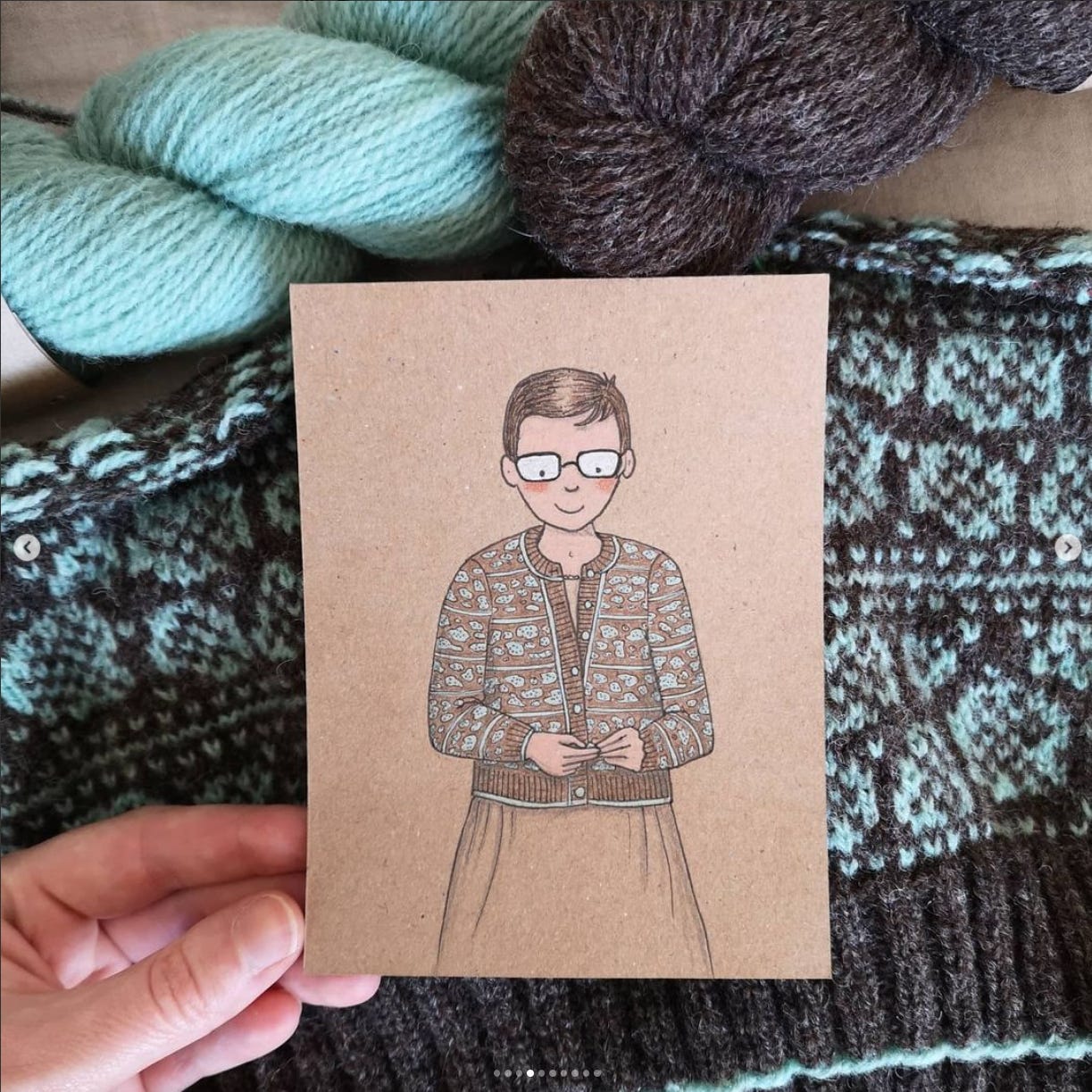 A white person's hand holds a sketch on a piece of brown paper, in front of a pile of knitting and yarn. The sketch is of a short-haired person with glasses buttoning up a colourwork cardigan. The cardigan is brown and pale blue with motifs that resemble lichen-covered tree bark. The knitting beneath the picture is said cardigan coming to life, with two skeins of yarn lying beside it.