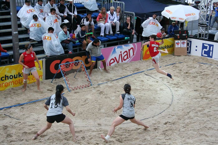 A sandy beach court with Mel on the right jumping in the air wearing a red muscle vest with Hibberd and 6 on and the shortest shorts ever. She's about to blast the ball at a trampoline at a 45 degree angle. There are two taiwanese players poised on a semi circle waiting to catch the ball.