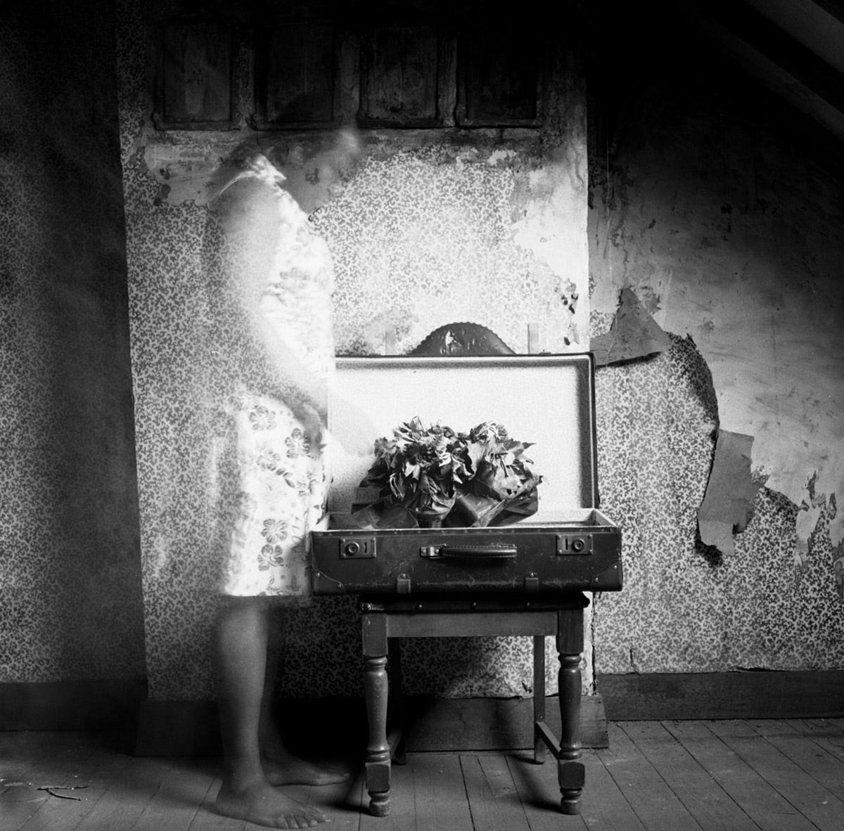 Black and white photo which is an autoportait of Hélène Amouzou, a Togolese artist, who uses this project to show her precarity during the decades long journey in obtaining her Belgian citizenship. In the photo she appears barefoot and ghost-like in a floral dress, in front of an open suitcase, in a sparsely decorated room with no carpet.
