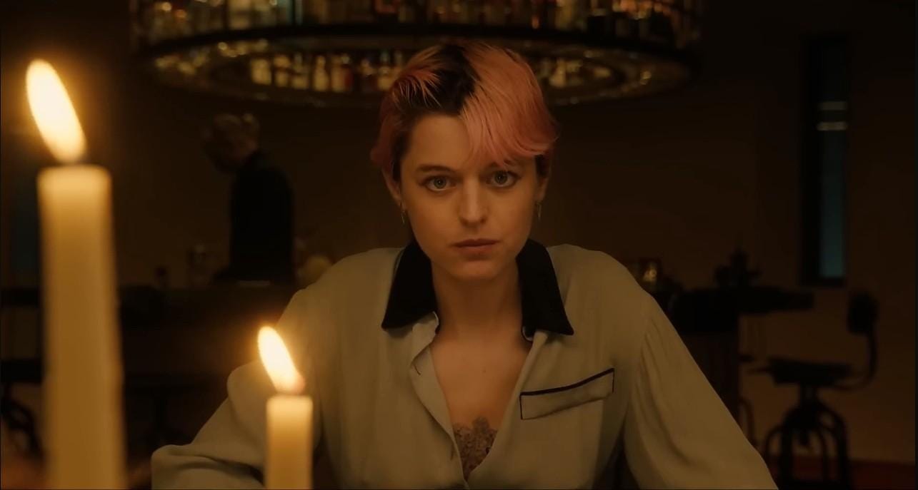 Actor Emma Corrin, a white nonbinary person with short pink hair, in a scene from A Murder At The End Of The World. They sit at a table staring into the camera, with lit candles burning in the foreground.