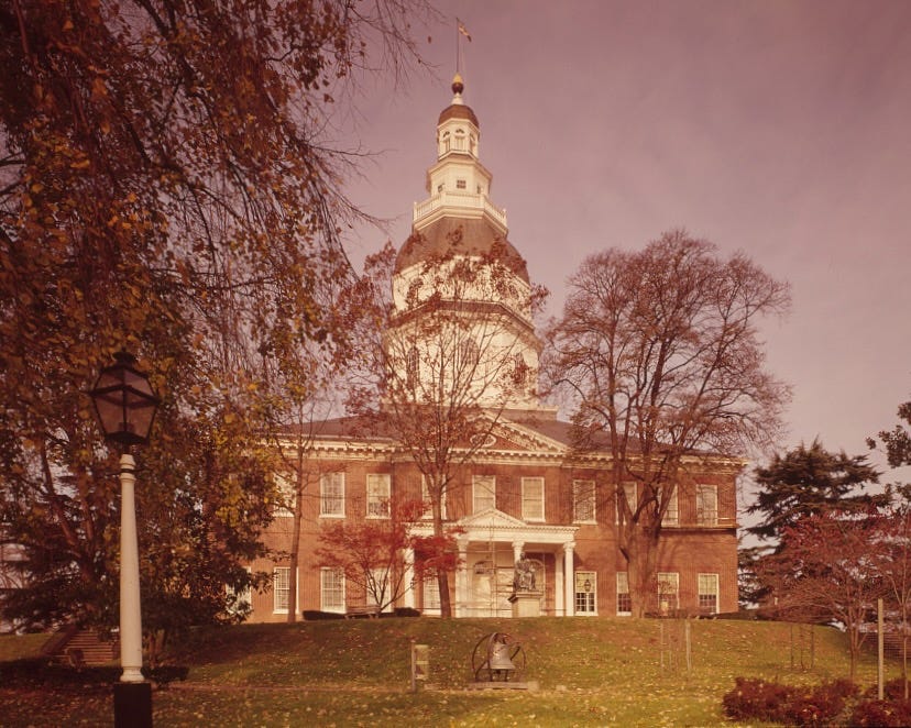 A slightly pink sepia-toned photograph of the Maryland State House sitting atop a hill with trees and a bell in the foreground. The base of the building is made mostly of brick.