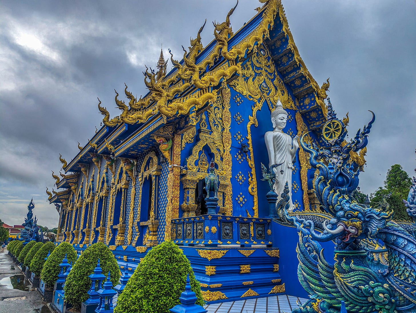 The ornately decorated Blue Temple which has lots of gold trim and other decorations. A white Buddha stands in front, a dragon guarding the entrance. 