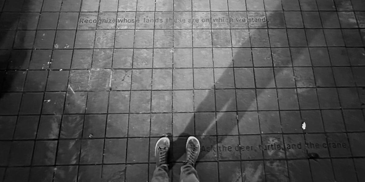 Person stands on sidewalk in a pair of lace-up sneakers. Imprinted in the concrete are the words, "Recognize whose lands these are on which we stand... Ask the deer, turtle, and the crane."