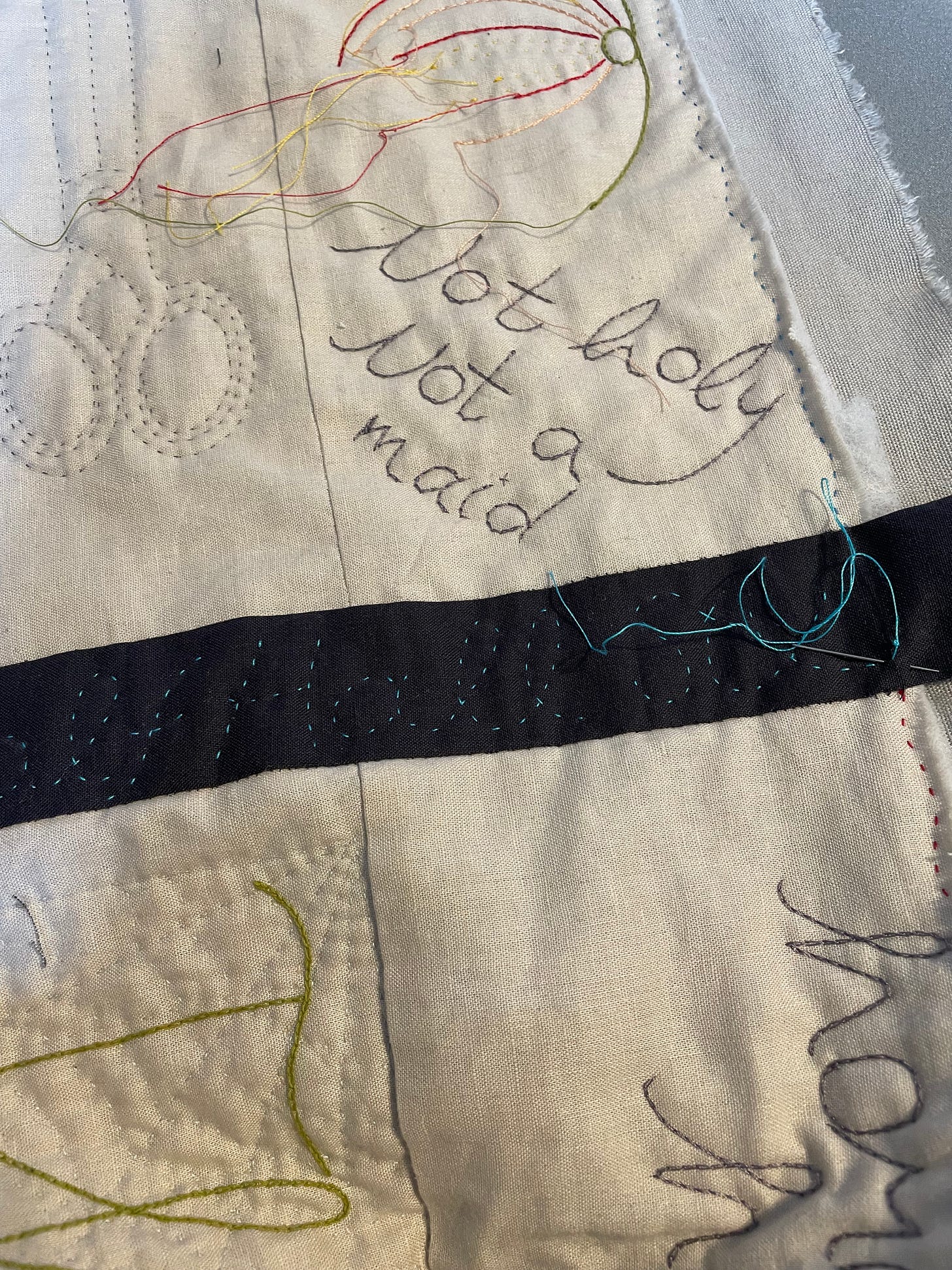 Quilted work - the words “Not holy Not a maid”, some scissors and a pomegranate are visible. There is a needle with blue thread in a strip of dark cloth.