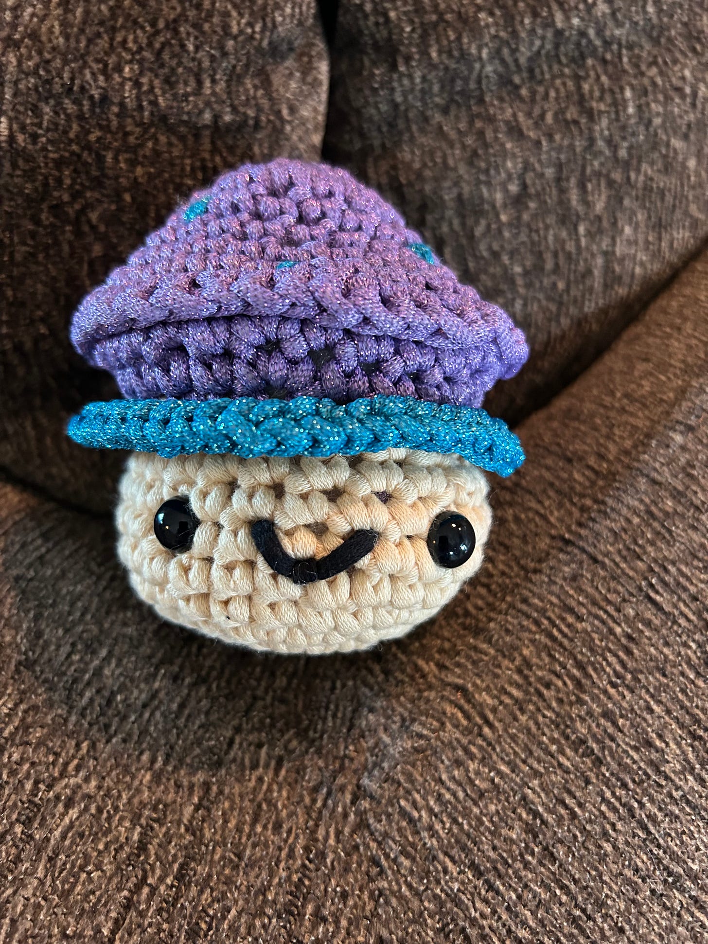 Crocheted mushroom with a purple and blue cap and tan stem with a smiley face.