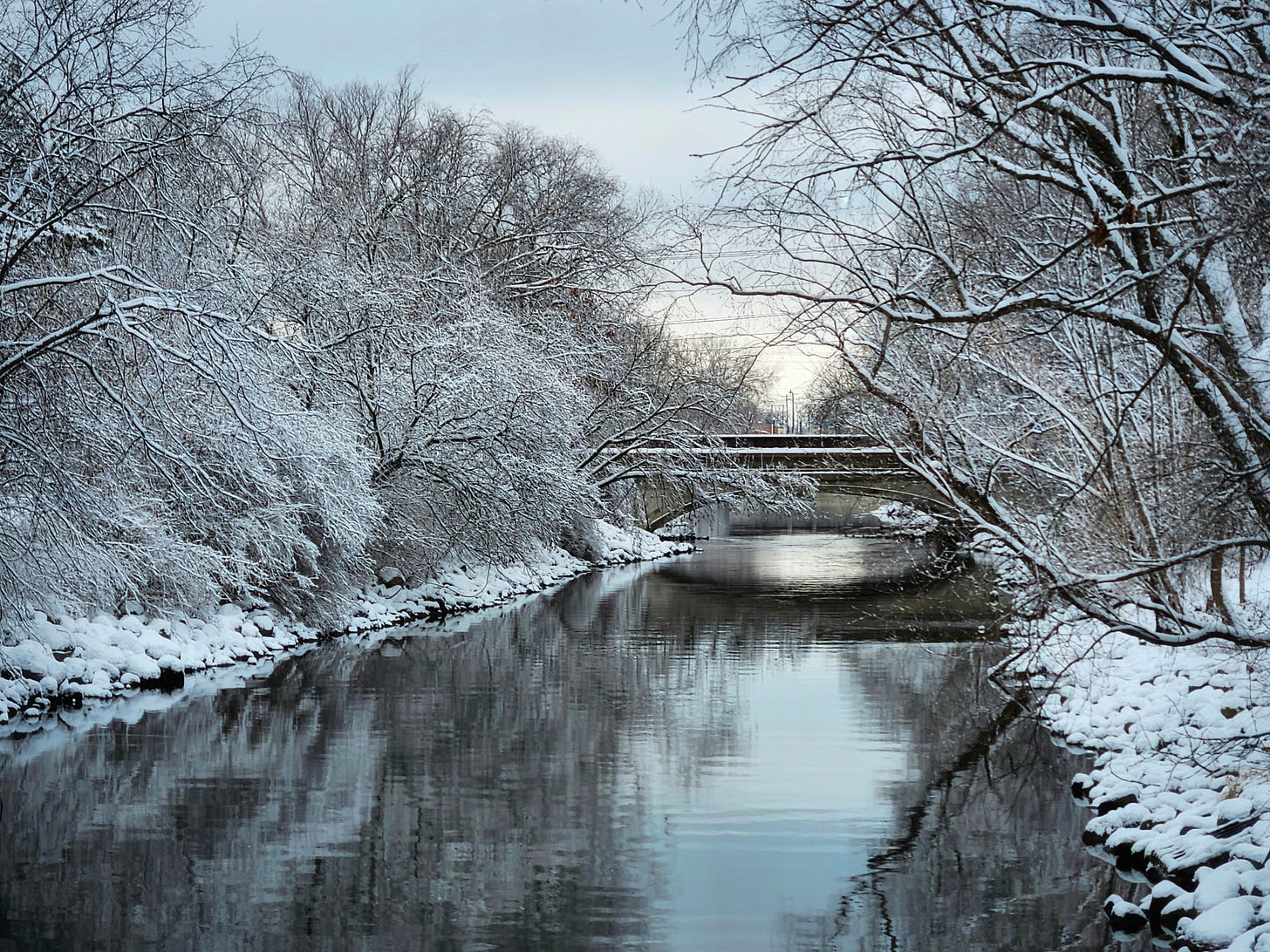 Black, mostly smooth water of a narrow river flows between snow-covered, rocky stream banks. Trees curve over the waterway, also covered in fluffy white snow. A bridge spans the water in the distance.