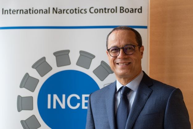 A man in a suit, wearing glasses, in front a poster that reads "International Narcotics Control Board" and "INCB"