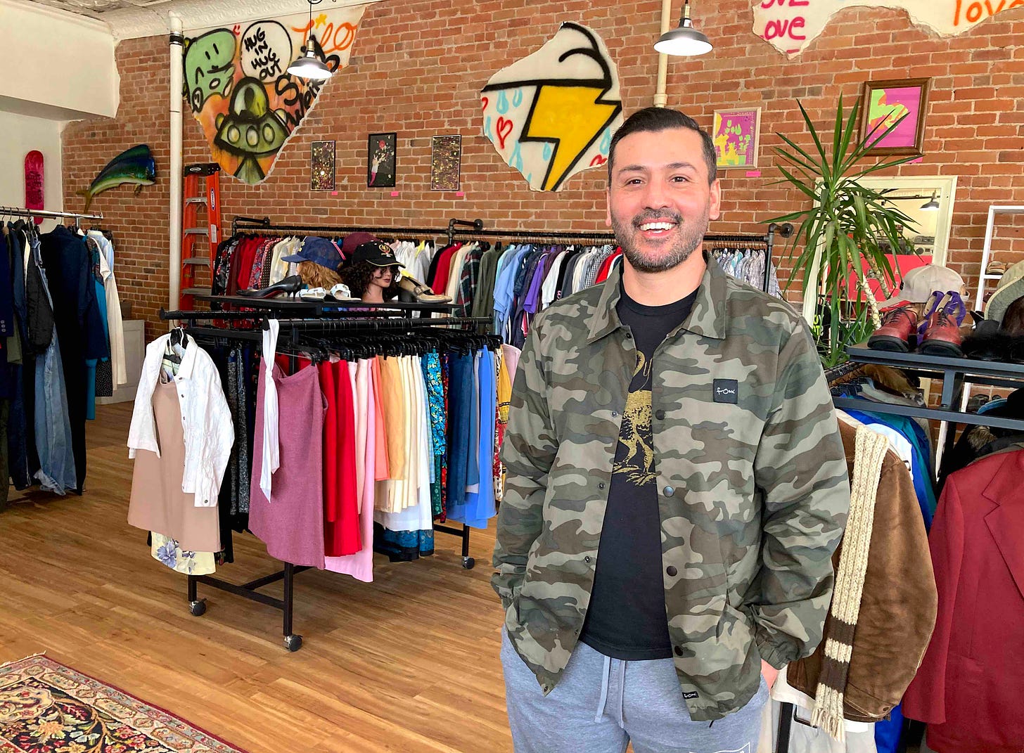 smiling man in green camo jacket and grey sweats in vibrant room full of clothing racks and exposed brick wall