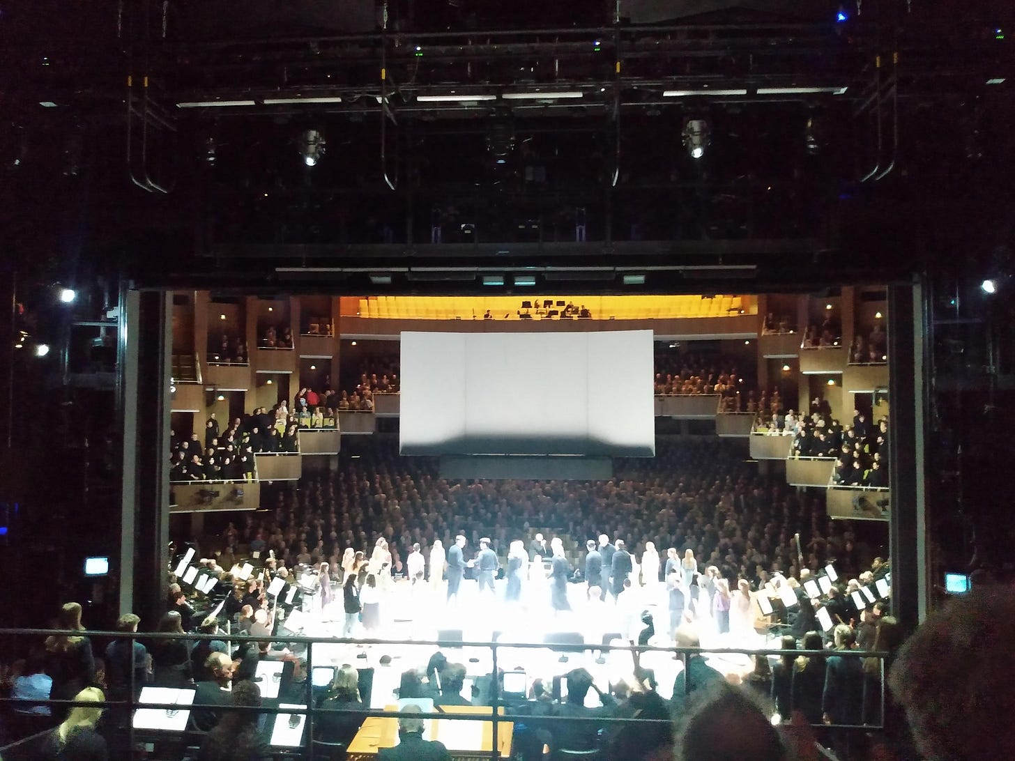 A central staged viewed from up-stage. Musicians and performers are on central stage receiving standing ovation from the audience.