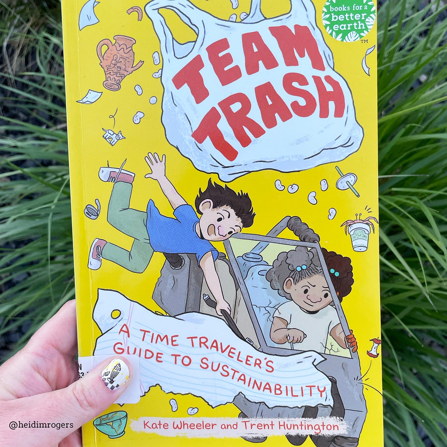 White woman's hand holding the young graphic novel, Team Trash, over long grass.