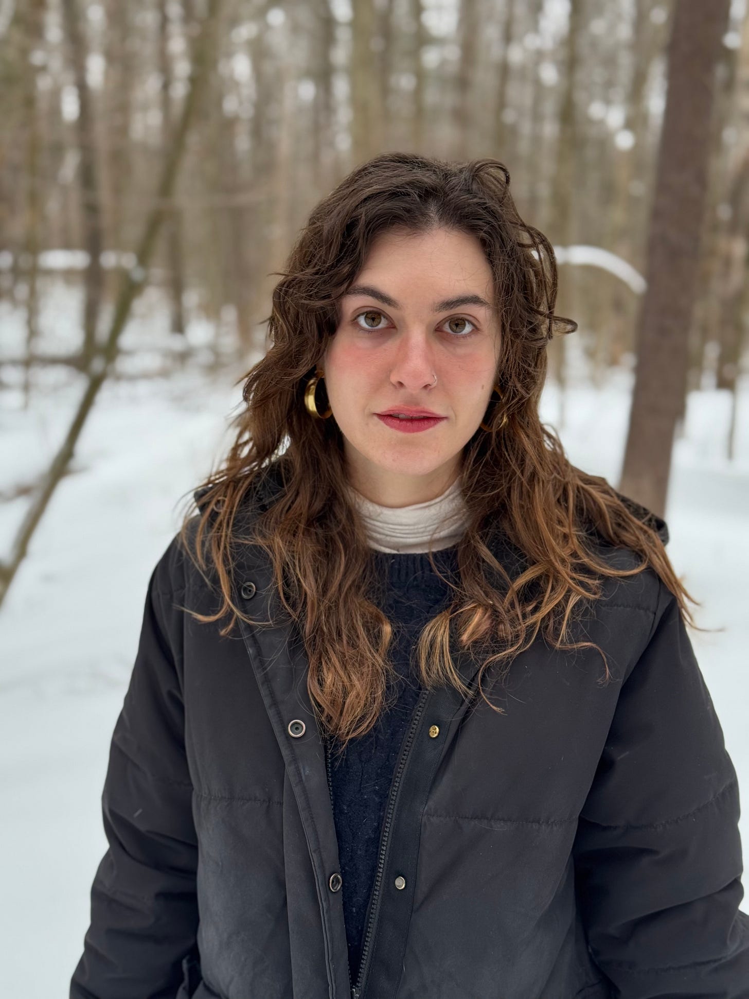 A woman with wavy brown hair and gold hoop earrings stands in a snowy forest, wearing a black puffer jacket and a layered sweater. She gazes directly at the camera with a calm expression.
