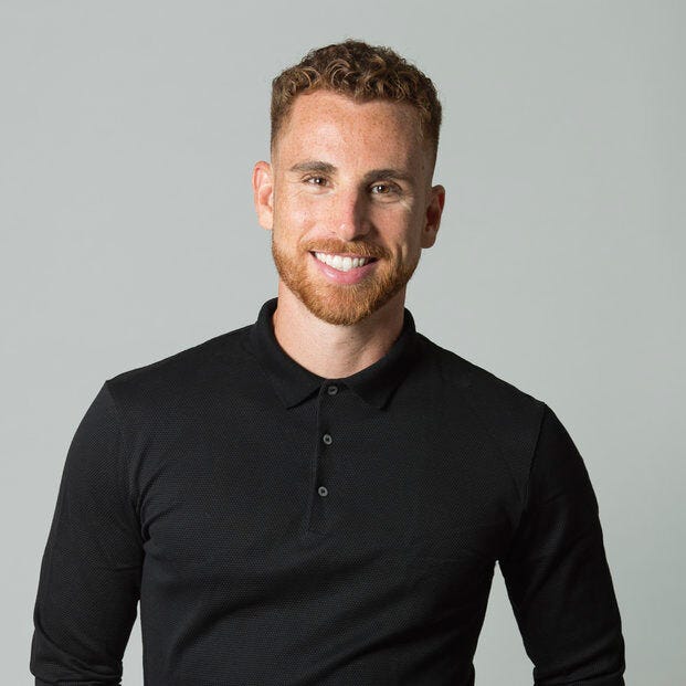 A person with short curly hair and a beard is smiling at the camera, exuding a friendly demeanor. They're donning a black long-sleeve shirt and standing against a plain light grey background—similar to what you'd see in professional portraits often showcased on gallery walls.