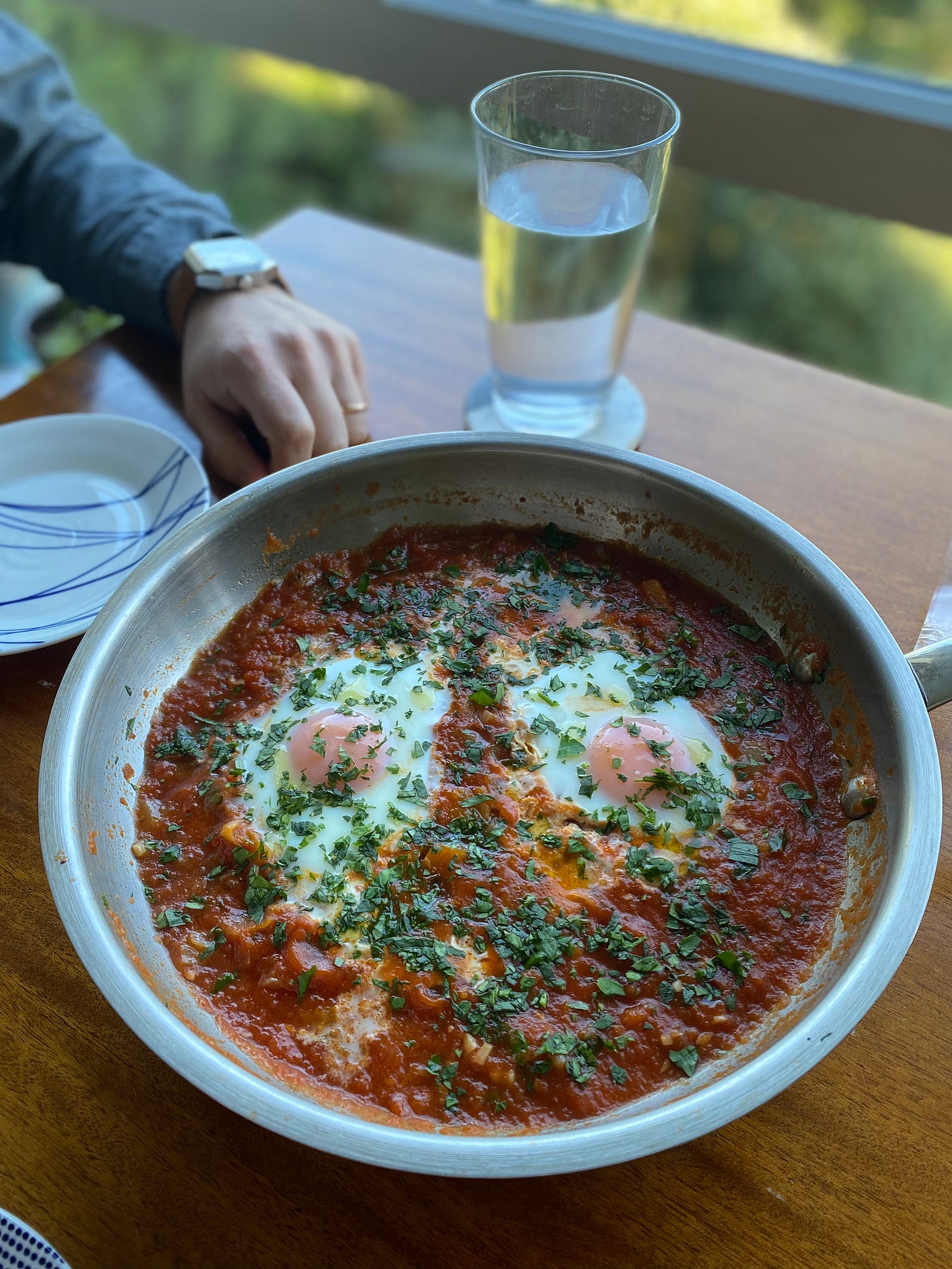 A deep red shakshuka in a steel pan with two eggs, sprinkled with parsley and drizzled with olive oil. Jeff's hand is on the table behind the pan.