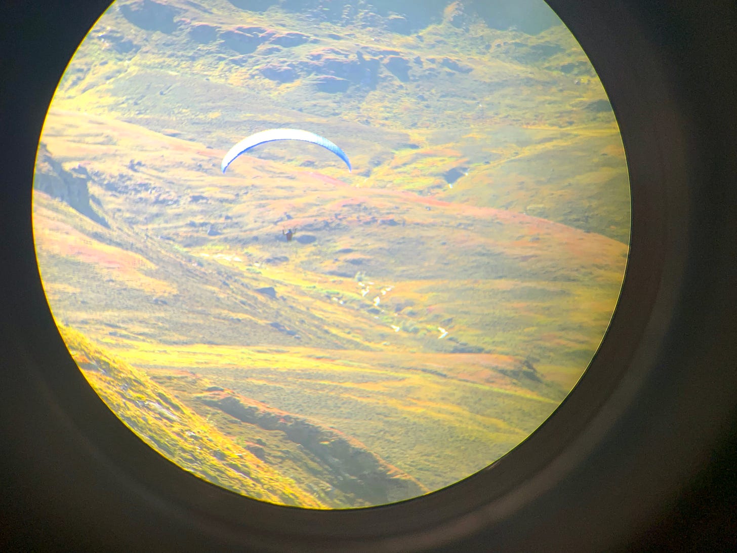 A hang-glider over grassy mountains, seen through a binocular lens.