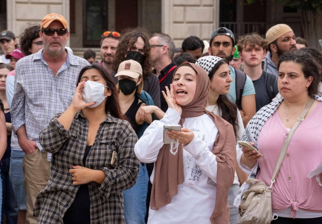 A woman in a hijab appears to shout and read from a phone in a crowd of people, one of them masked, another a woman wearing a hijab