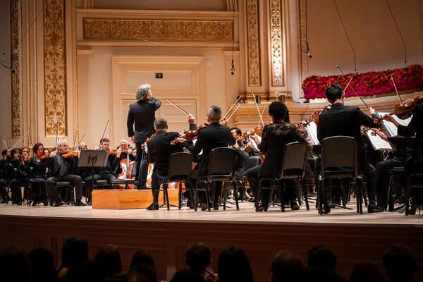 On the stage of Carnegie Hall, we see the conductor Gustavo Dudamel from the back, surrounded by string players. 