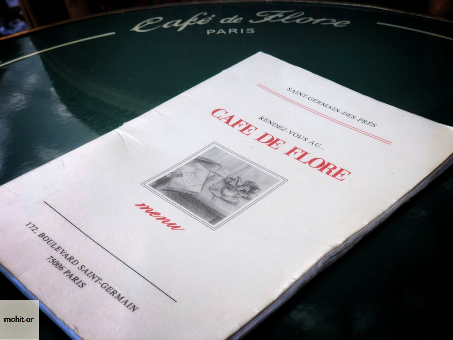 A menu from Café de Flore in Paris, resting on a dark green table surface. The white menu cover features the restaurant's name in red text, along with "SAINT-GERMAIN-DES-PRÉS" at the top. There's a small black and white illustration in the center, and the address "172, BOULEVARD SAINT-GERMAIN 75006 PARIS" is printed at the bottom. The café's name is also visible embossed on the table surface in the background.