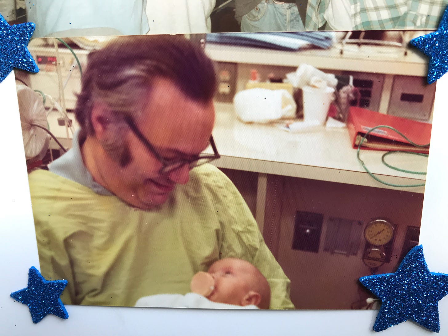 A bespectacled, middle-aged man with swept-back black-and-gray hair and sideburns, and wearing a green hospital smock, holds a tiny baby as he sits in a hospital ward. The baby, sucking on a pacifier, gazes back up into his father's eyes.