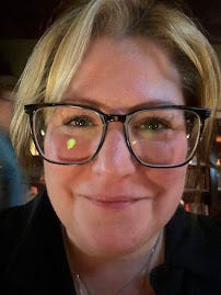 A close up of me, in a dark room (bar), with large black-framed glasses, grinning into the camera. I've got a blonde fringe and the rest of my hair it tied back. I'm wearing a black shirt.