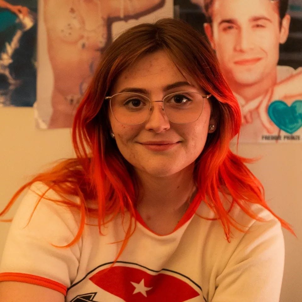 Hannah, a person with brown hair with orange tips, glasses, and a white shirt smiles at the camera