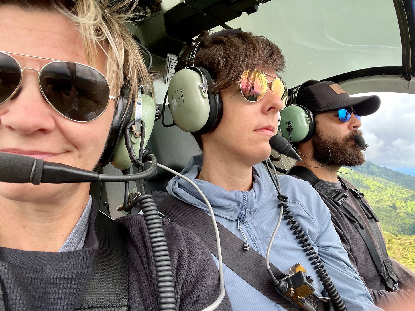 Two women and one bearded man wear headsets as they fly over Hawaii in a doorless helicopter.