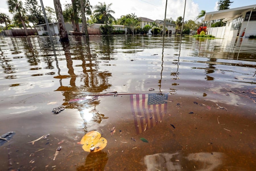 A small American flag sits in floodwater, near houses.