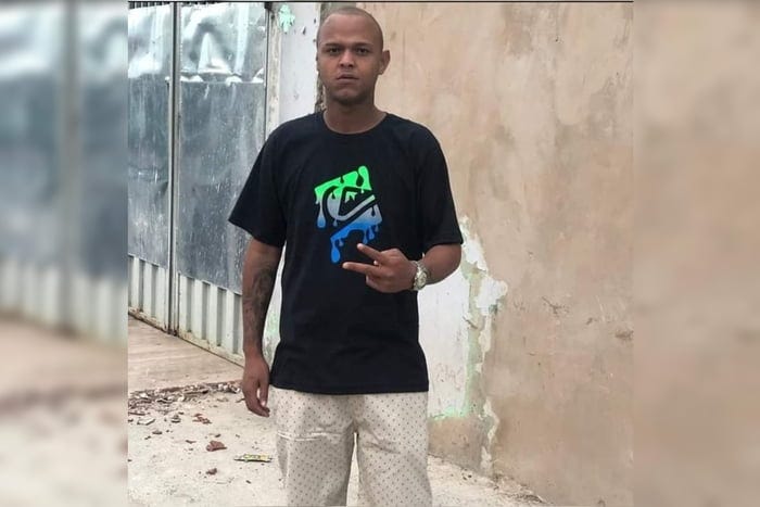 Imagem colorida de um homem negro, de camiseta preta e bermuda bege. Ele faz um sinal de cumprimento com a mão esquerda, à altura da barriga