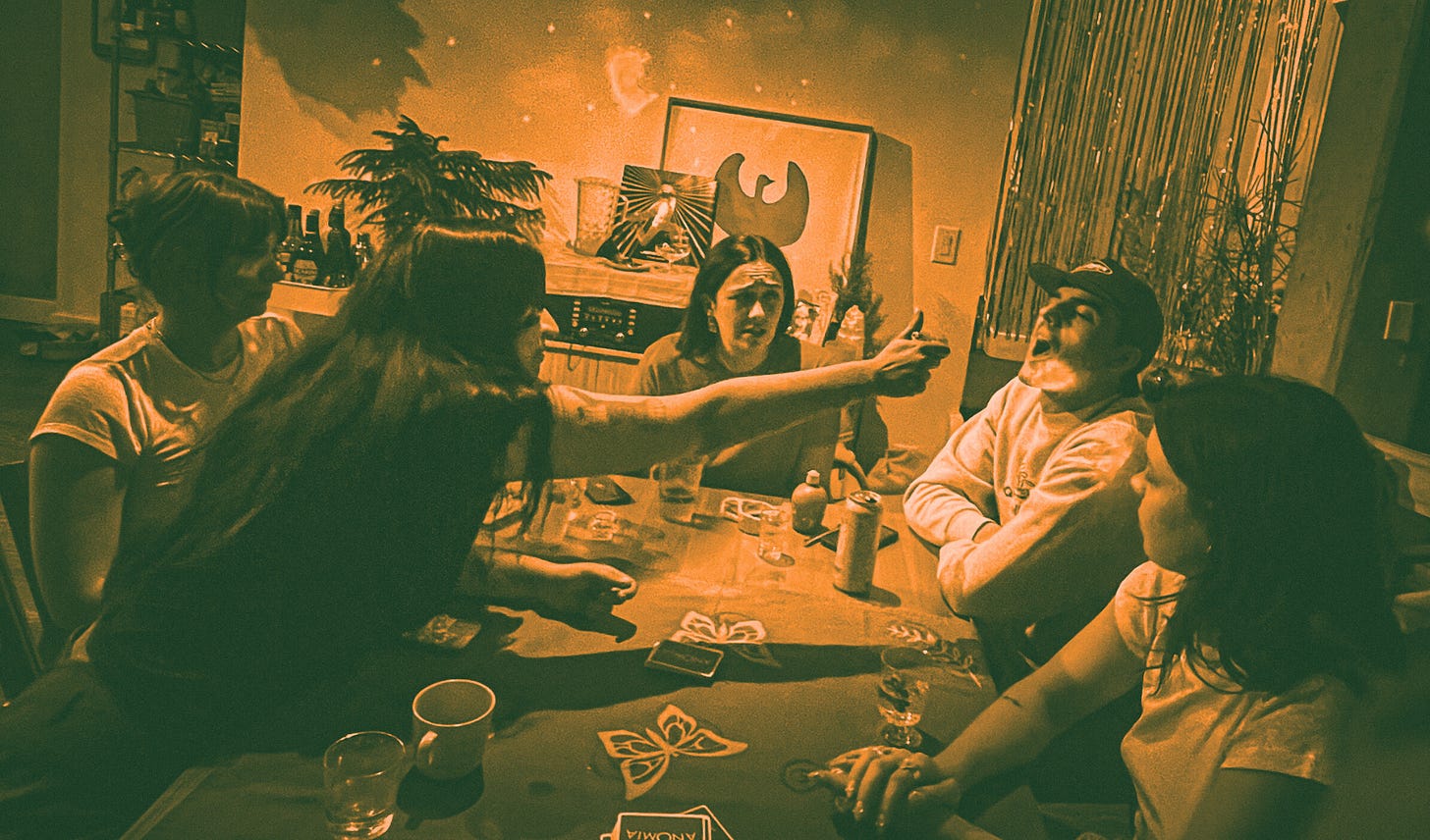 five people sitting around a table while one reaches across the table and shoots the content of a mini water gun into anothers mouth. Anomia cards are on the table.