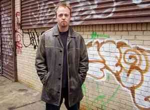 Photo of a weary-looking undercover cop standing in front of a brick wall covered in graffiti.