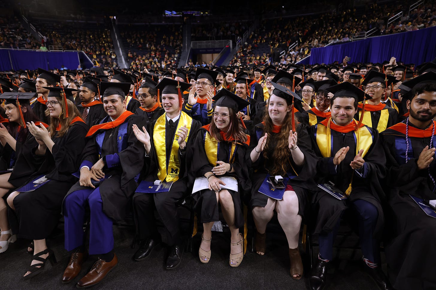 A row of graduates applaud and smile as a large crowd of other graduates fills the stands of a basketball arena.