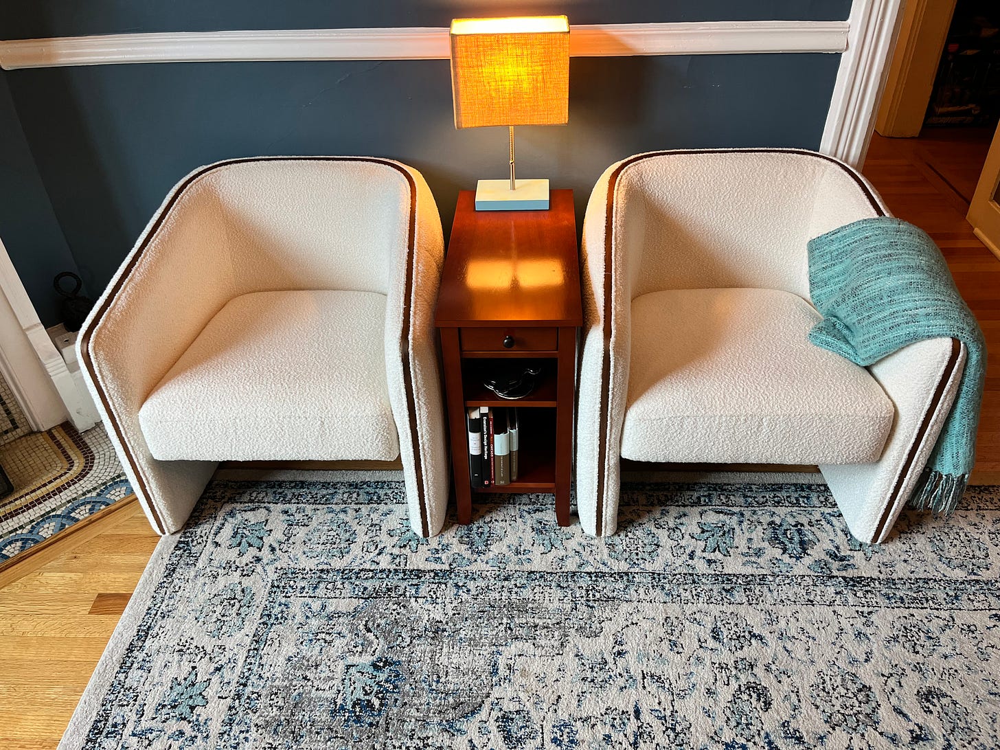 Two white chairs against a blue painted wall. A light blue throw blanket rests on one, and a blue and white rug sits in front of them. 