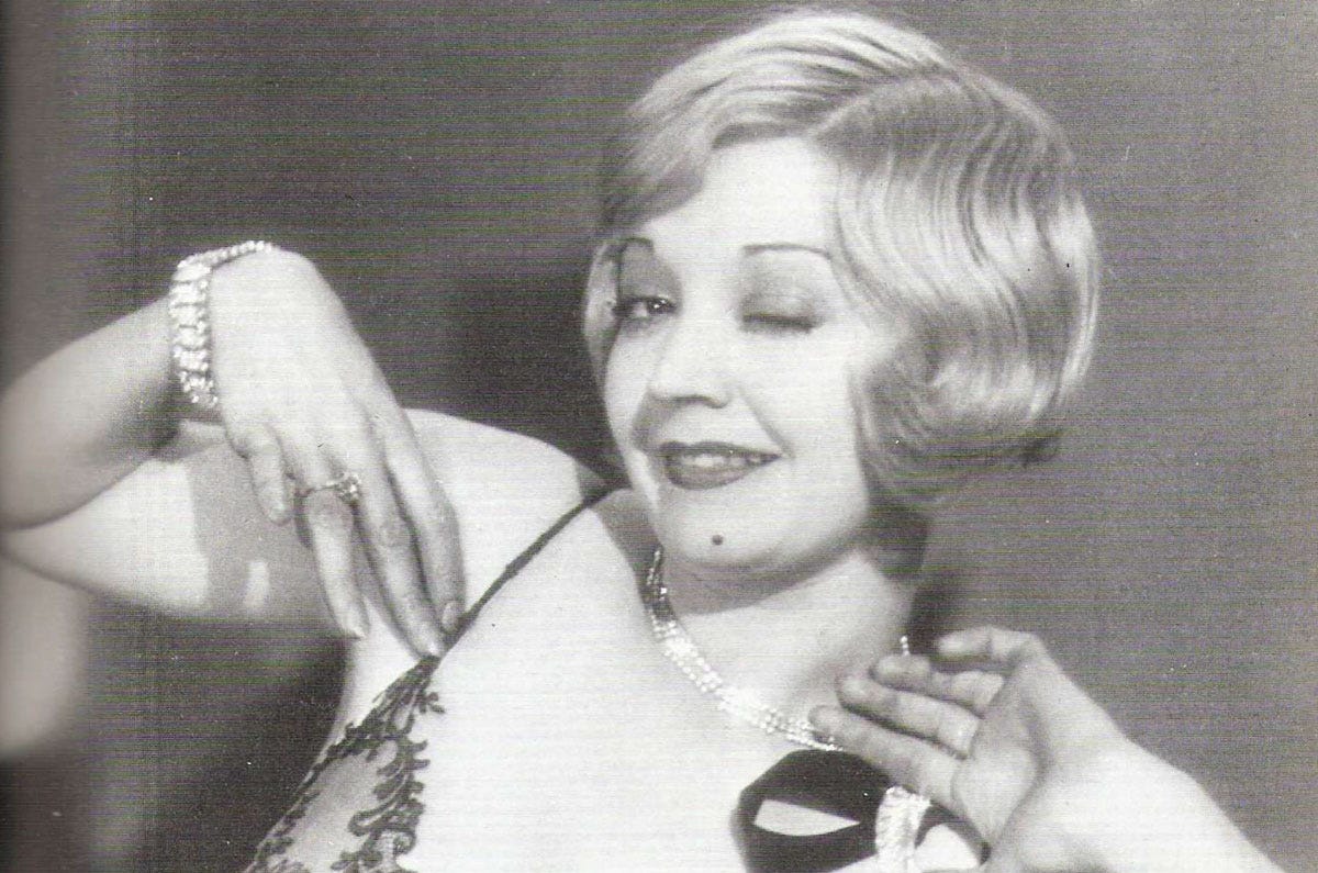 Woman in an evening dress, with bleached blonde hair and jewellery winks at the camera
