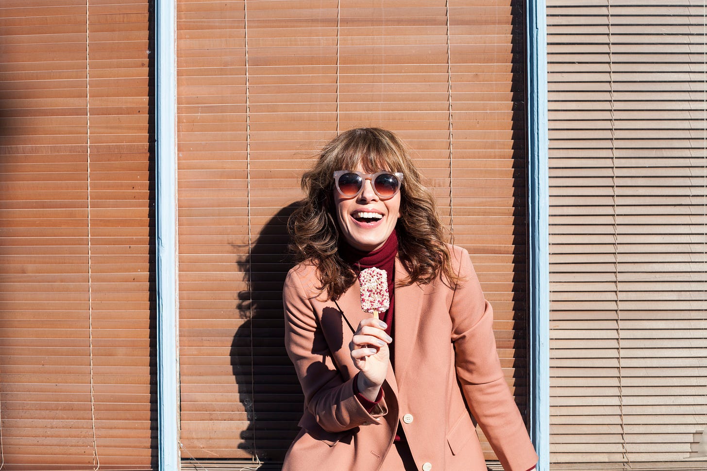 A woman with dark blonde long curly hair and bangs sits on a window sill holding an ice-cream. She is smiling and wearing pink sunglasses and a suit with a burgundy roll neck knit.
