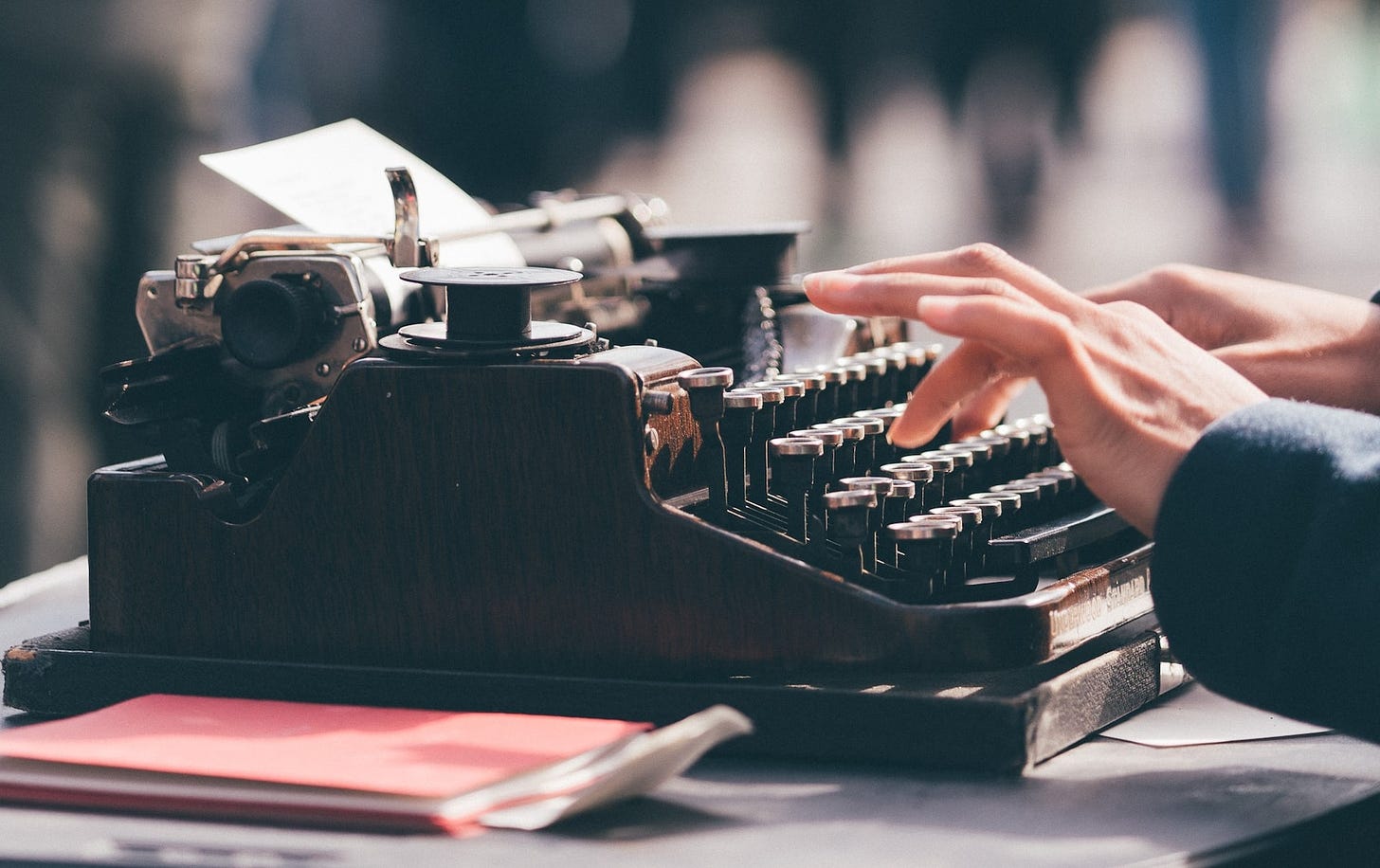 Photograph of hands typing on a vintage typewriter, anotebook alongside on the table