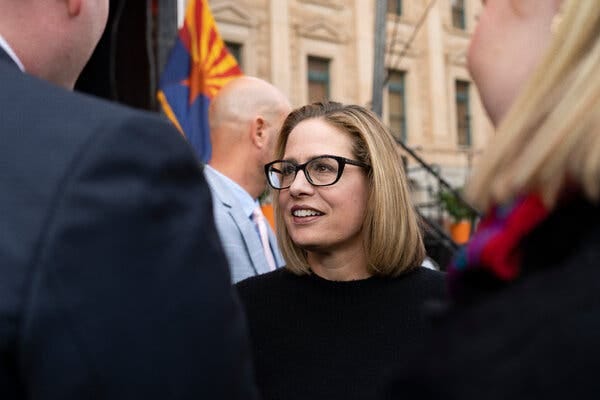 Kyrsten Sinema, wearing a black top with black glasses, talks to a small group of people. There is an Arizona state flag in the background.