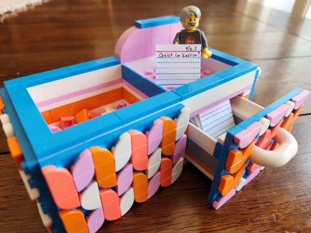 What appears to be a small two drawer zettelkasten made out of LEGO in a somewhat garish blue, white, orange and pink color scheme. The right drawer is open with a few dozen mini-index cards inside. On top is a LEGO figurine with gray hair, a beard. and mustache. It is holding a tiny index card that reads "9/8,3 Geist im Kasten?"