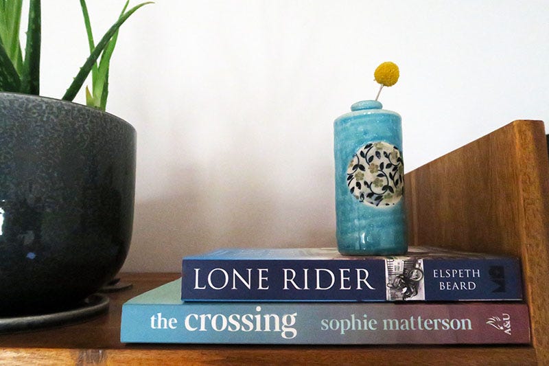 Two books, 'Lone Rider' by Elspeth Beard and 'The Crossing' by Sophie Matterson, stacked on a bookshelf with a small vase on top of them.