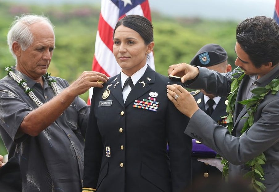 Tulsi Gabbard being decorated in her military uniform by two individuals, standing proudly in front of the American flag.