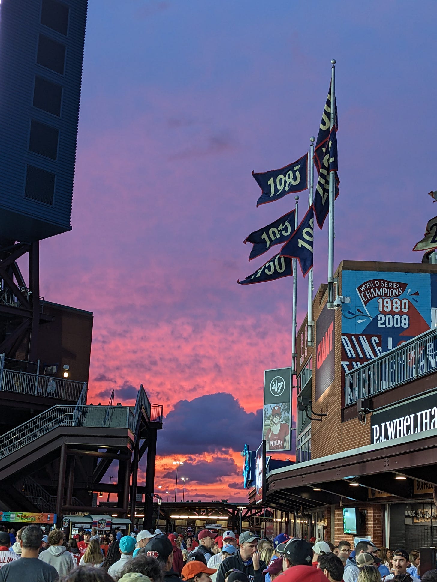 A dramatic sunset, fading from purple-blue to pink, is framed by rippling flags representing the years the Philadelphia Phillies won the pennant. In the foreground, a crowd of baseball fans.