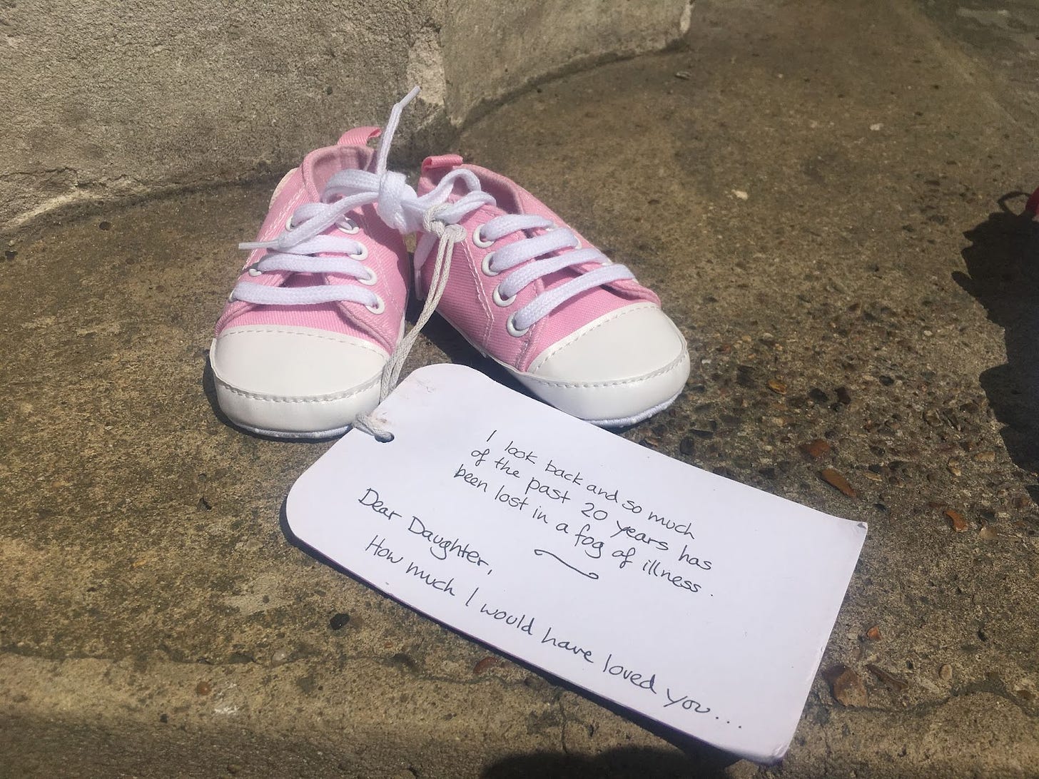 Photo from the Millions Missing action for ME/CFS in 2019. Image description: pink, baby-sized Converse trainers with a handwritten tag that reads: “I look back and so much of the past 20 years has been lost in a fog of illness. Dear Daughter, how much I would have loved you…”.